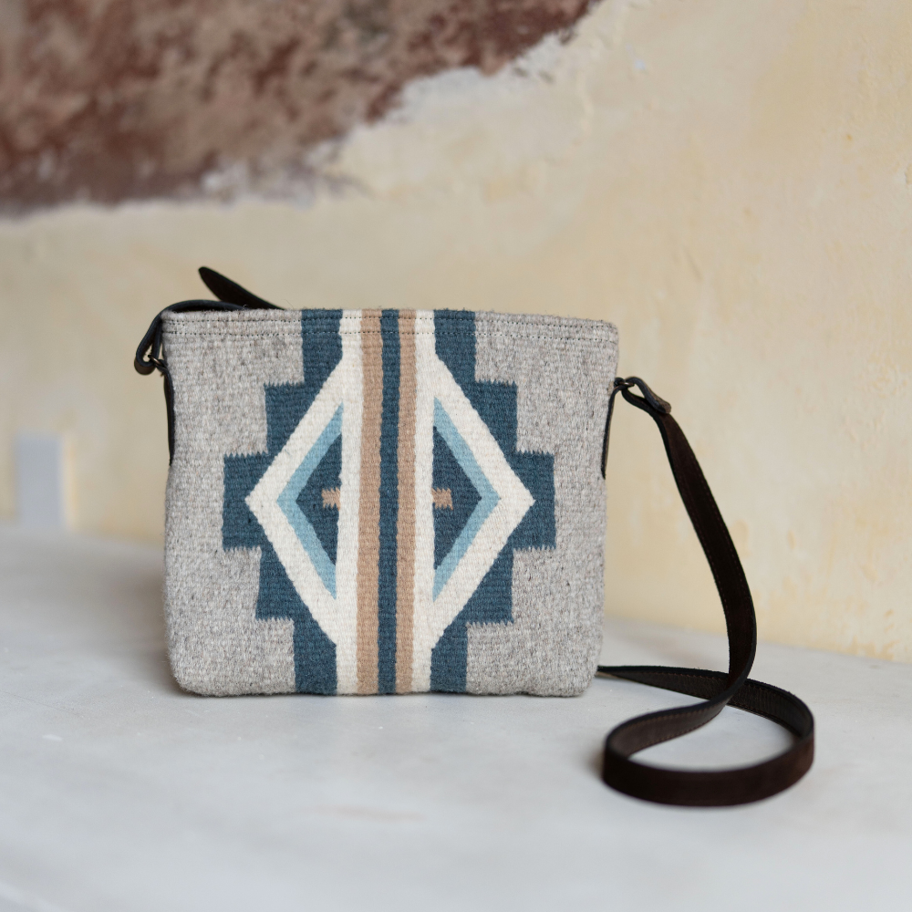 AMASOUK Sueños Crossbody with dark brown leather strap and handwoven wool in blue, heather gray, and ivory. Features traditional Zapotec patterns and artisan craftsmanship.