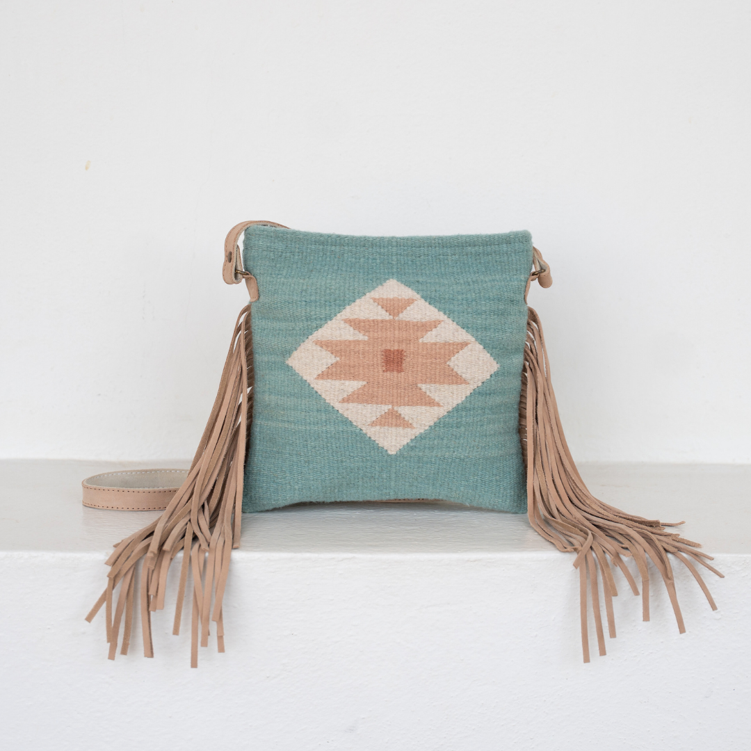 Handwoven fringe crossbody bag with abstract Zapotec butterfly design in soft blue tones; features flowing fringe, adjustable leather strap, and two internal pockets