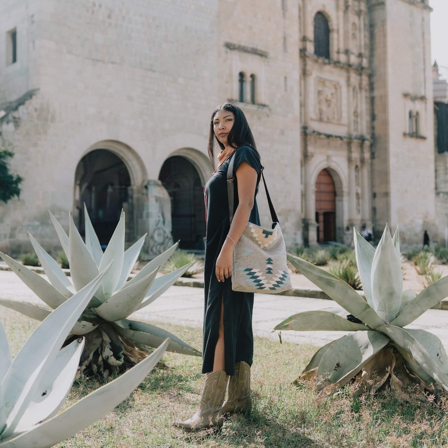 Fashionable woman with an AMASOUK handwoven wool Aguila Shoulder/Crossbody bag in a Zapotec design with blue, ivory, and honey on a gray background. Ideal for day-to-night versatility.