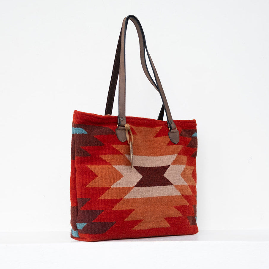 A woven tote bag with a Southwestern geometric pattern in red, orange, brown, and beige, featuring dark brown leather shoulder straps, set against a plain white background.