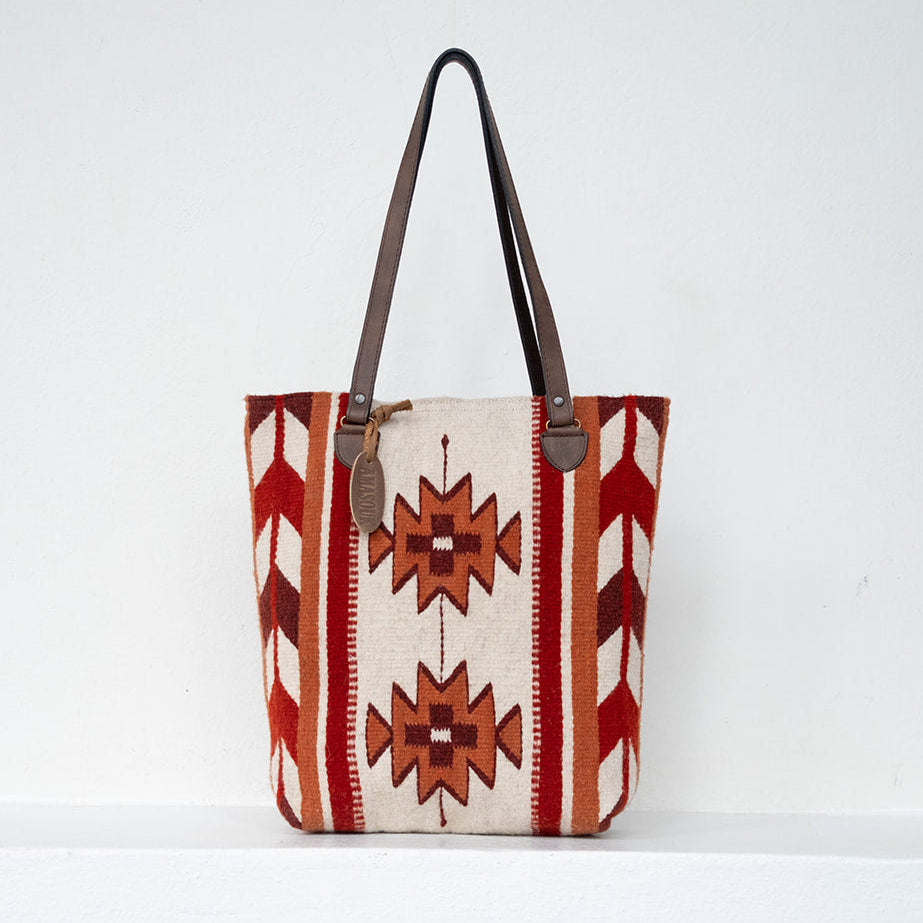 A woven tote bag with a Southwestern geometric pattern in red, orange, and cream, featuring dark brown leather shoulder straps, set against a plain white background.