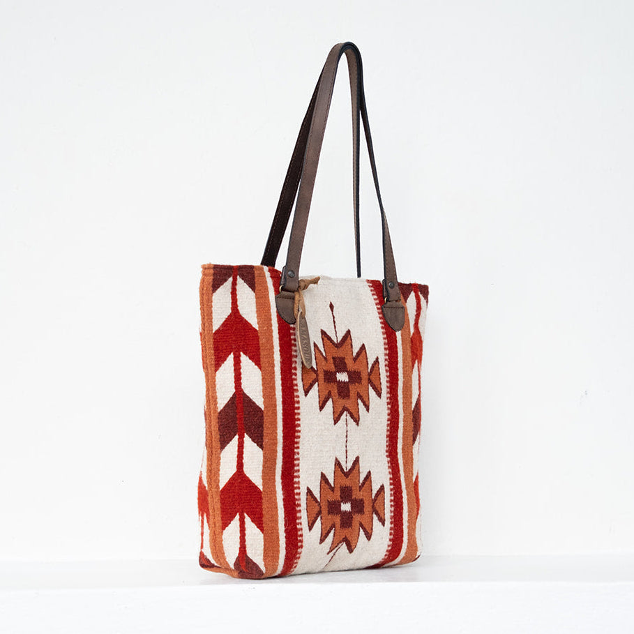 A Southwestern-style woven tote bag with geometric patterns in rust red, orange, and brown on a cream background, featuring dark brown shoulder straps, set against a plain white studio background.