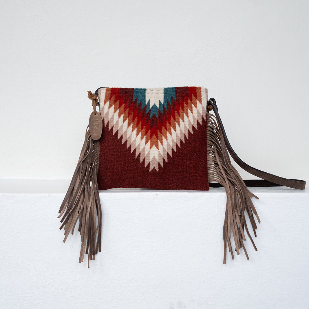 A small fringe clutch bag with a Southwestern-style woven chevron pattern in burgundy, cream, and teal, featuring brown faux-leather fringe tassels and a matching shoulder strap, displayed on a white ledge against a white wall.