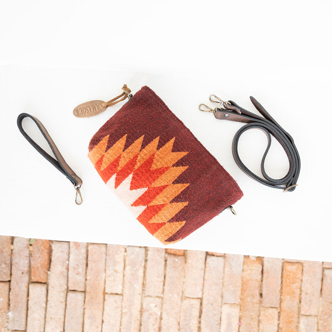 A woven clutch bag with a Southwestern-style geometric pattern in rust red, orange, and cream, shown with a black wristlet strap and a black shoulder strap on a white surface with a brick-patterned border at the bottom.