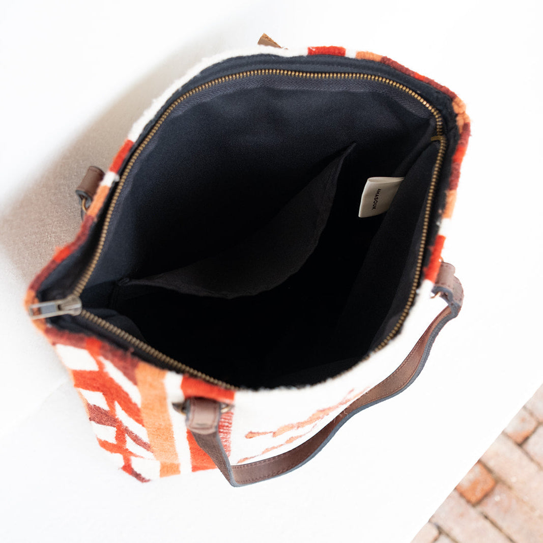 An open top view of a patterned shoulder bag with a black interior lining, featuring orange, white, and brown geometric stripes, resting on a white surface with a brick floor visible in the corner.