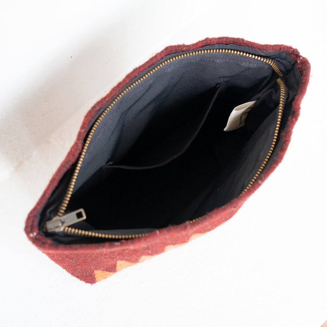 An open zippered pouch or clutch shown from above on a white surface. The exterior fabric is a textured rust red or terracotta color with a subtle geometric pattern near the bottom edge, and the interior is lined in black with a gold zipper.