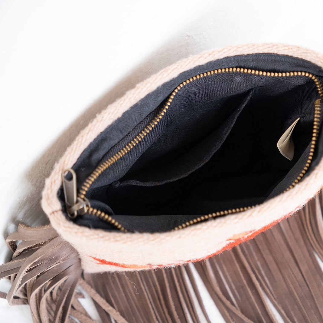A close-up of a fringed clutch bag with a cream-colored woven trim, taupe faux-leather fringe, a black interior lining, and gold-tone zipper hardware, shown on a white background.