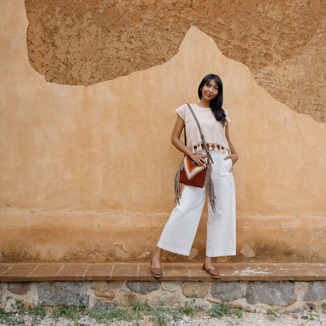 A woman stands against a textured ochre stucco wall wearing a white sleeveless top and wide-leg white pants, with a brown and red geometric patterned shoulder bag and brown sandals.