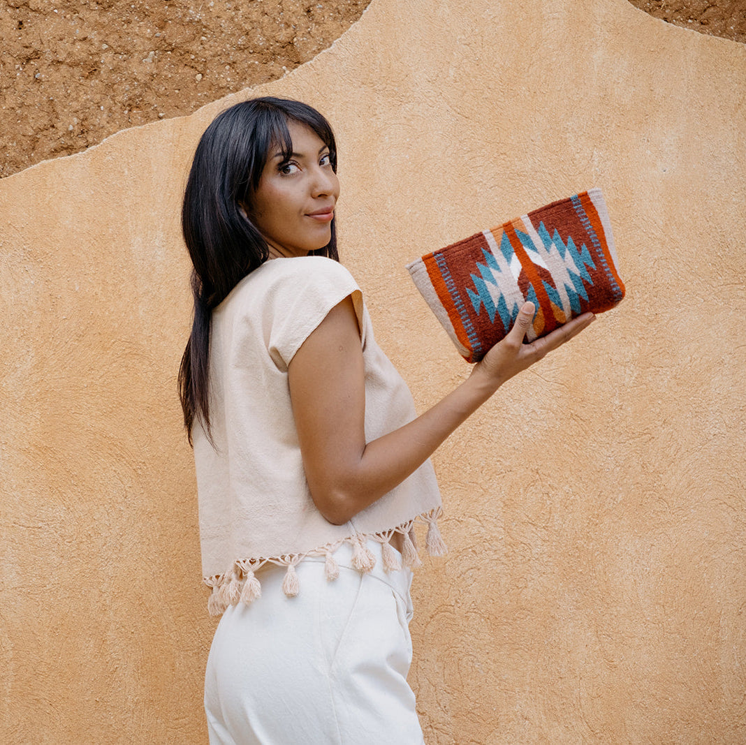 A woman with long dark hair stands against a textured beige wall, holding a woven clutch bag with a red, blue, and white geometric pattern.