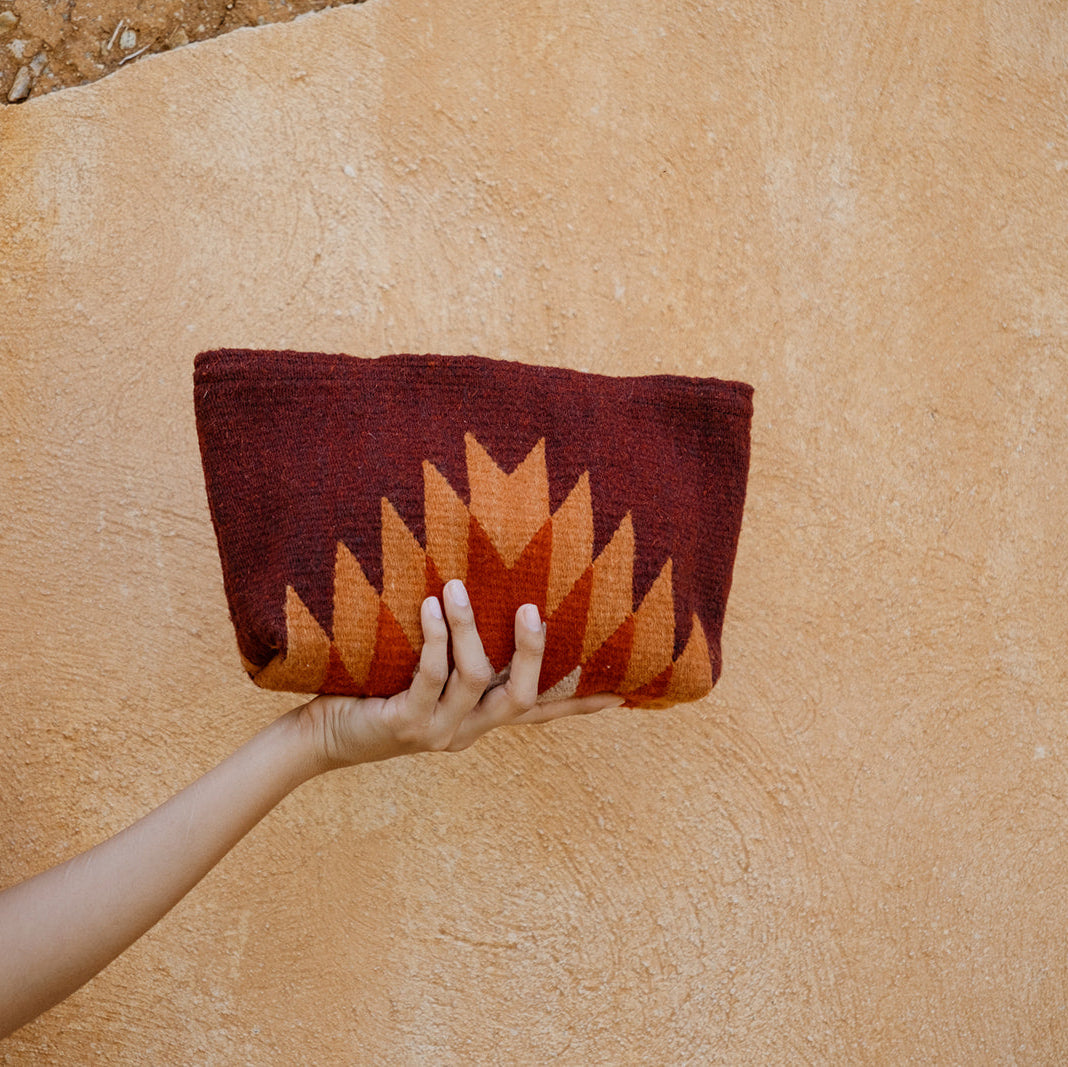 A person holds a small woven clutch bag with a geometric pattern in dark red, rust orange, and brown against a textured beige stucco wall.