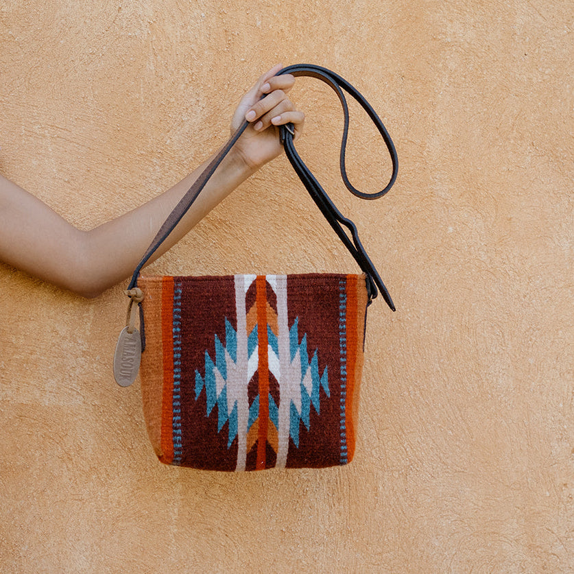 A woman in a light pink dress holds up a small woven shoulder bag with a Southwestern geometric pattern in brown, red, white, and blue, set against a textured beige stucco wall.