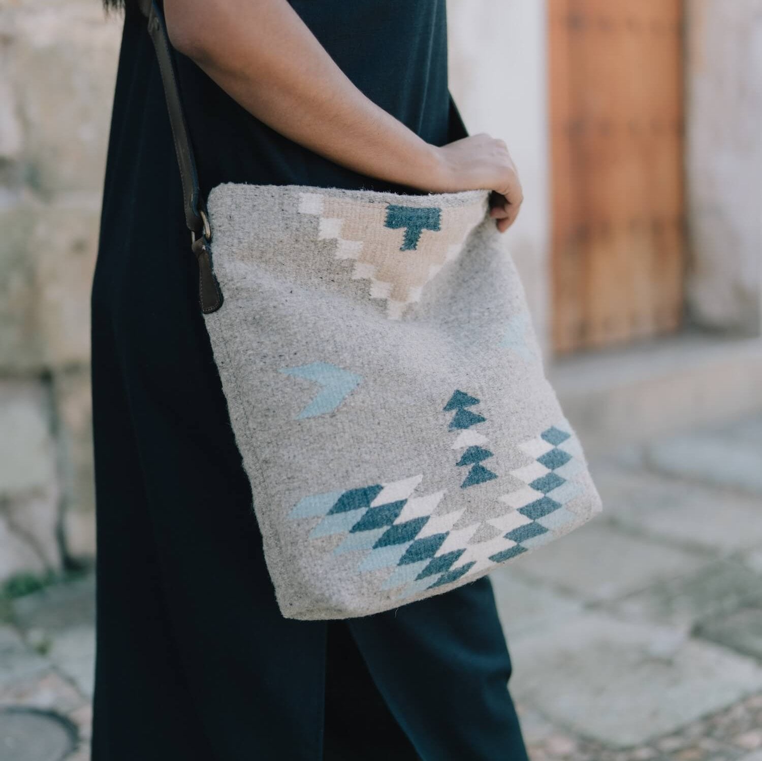 Woman with an AMASOUK handwoven wool Aguila Shoulder/Crossbody bag showcasing a Zapotec pattern in blue, ivory, and honey on a gray backdrop. Perfect for everyday activities and evening events.