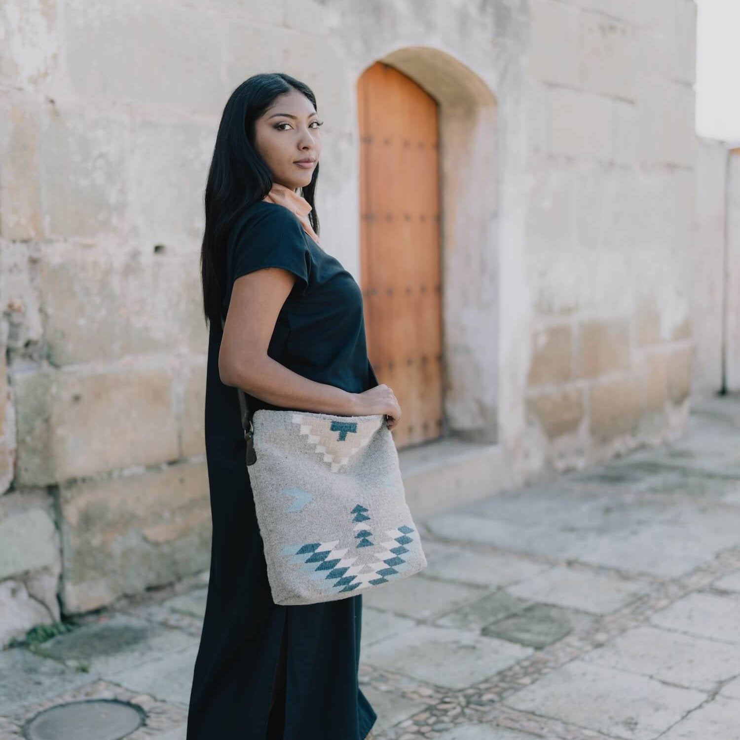 Effortlessly chic woman carrying an AMASOUK handwoven wool Aguila Shoulder/Crossbody bag with a Zapotec design in blue, ivory, and honey on a gray background. A versatile accessory for any occasion.