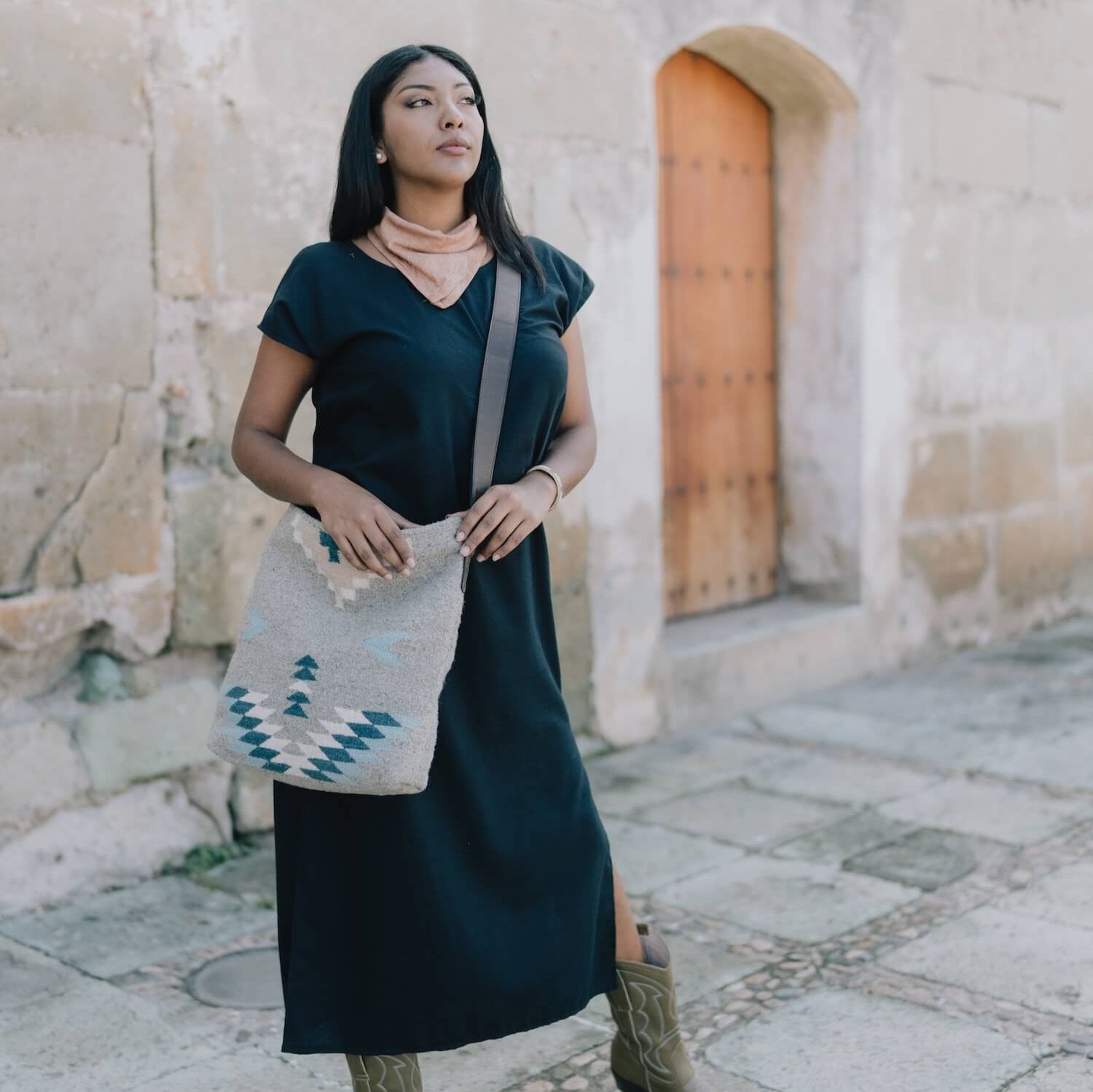 Woman carrying an AMASOUK handwoven wool Aguila Shoulder/Crossbody bag featuring a Zapotec pattern in blue, ivory, and honey on a gray backdrop. Perfect for errands, work, or casual outings.