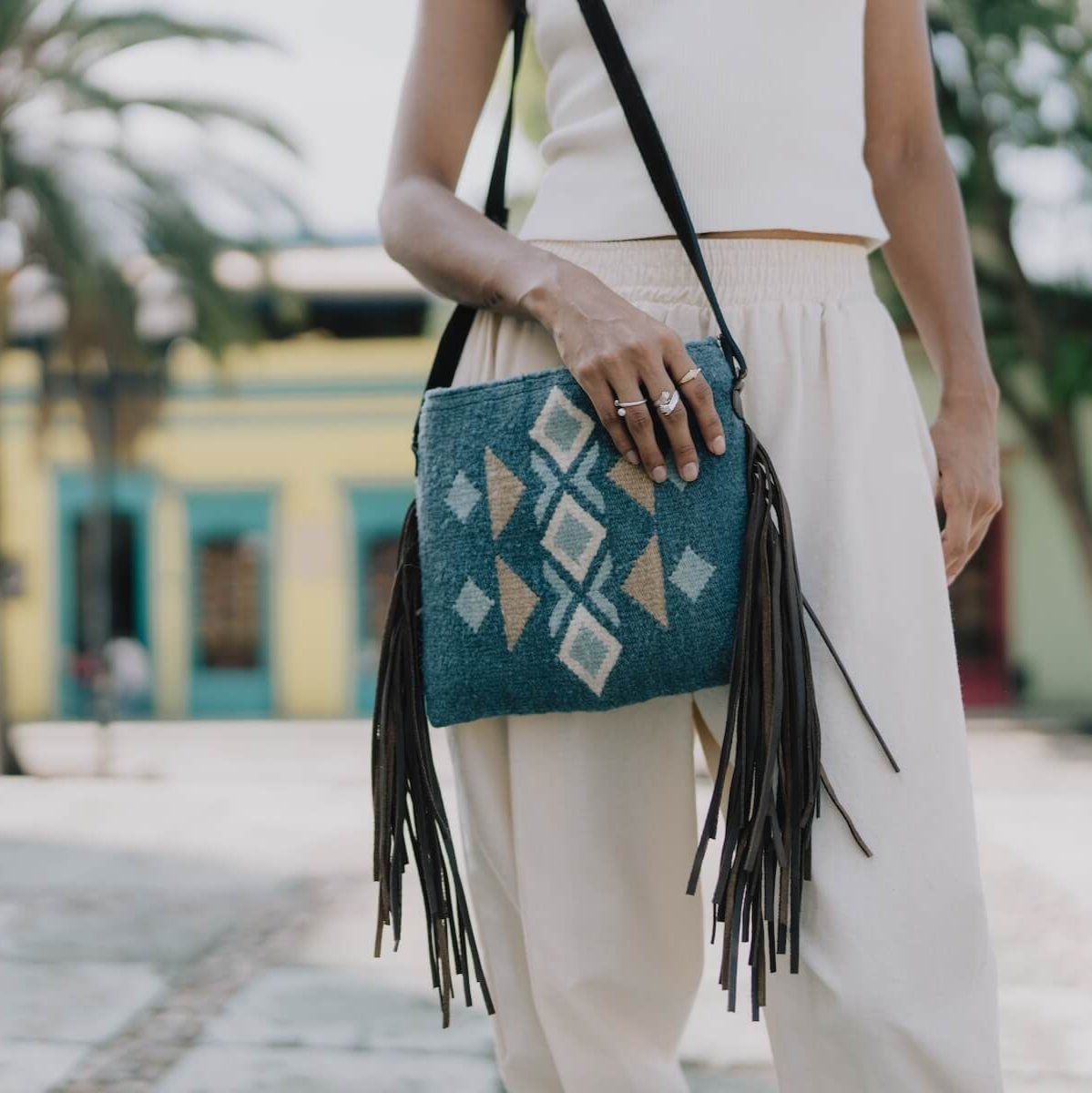 Woman carrying an AMASOUK Jalisco Fringe Crossbody bag with dark brown leather strap and fringe sides, featuring handwoven wool in blue, heather gray, and honey. Festive design with traditional Zapotec patterns.