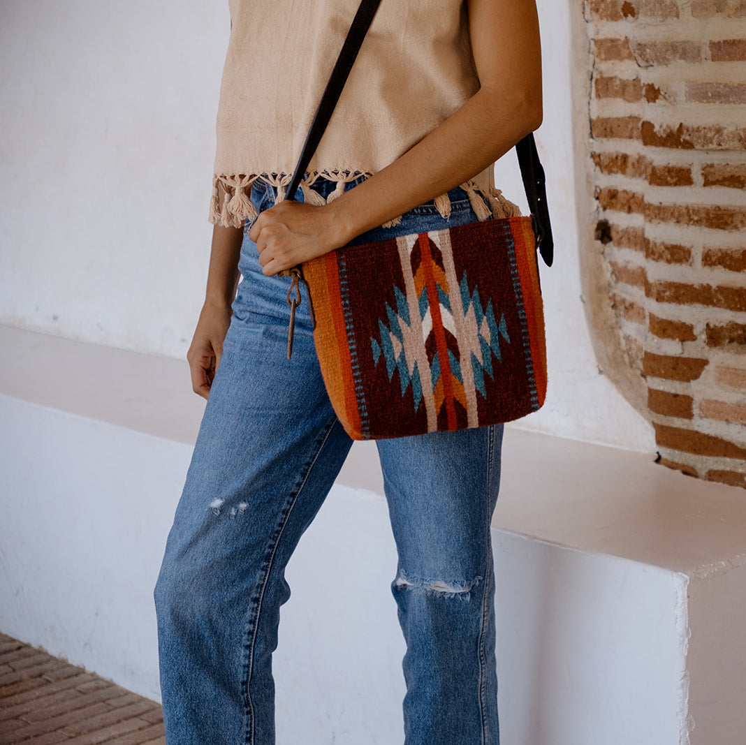 A person wearing blue jeans and a beige top carries a brown woven crossbody bag with a colorful geometric pattern featuring orange, blue, and white.
