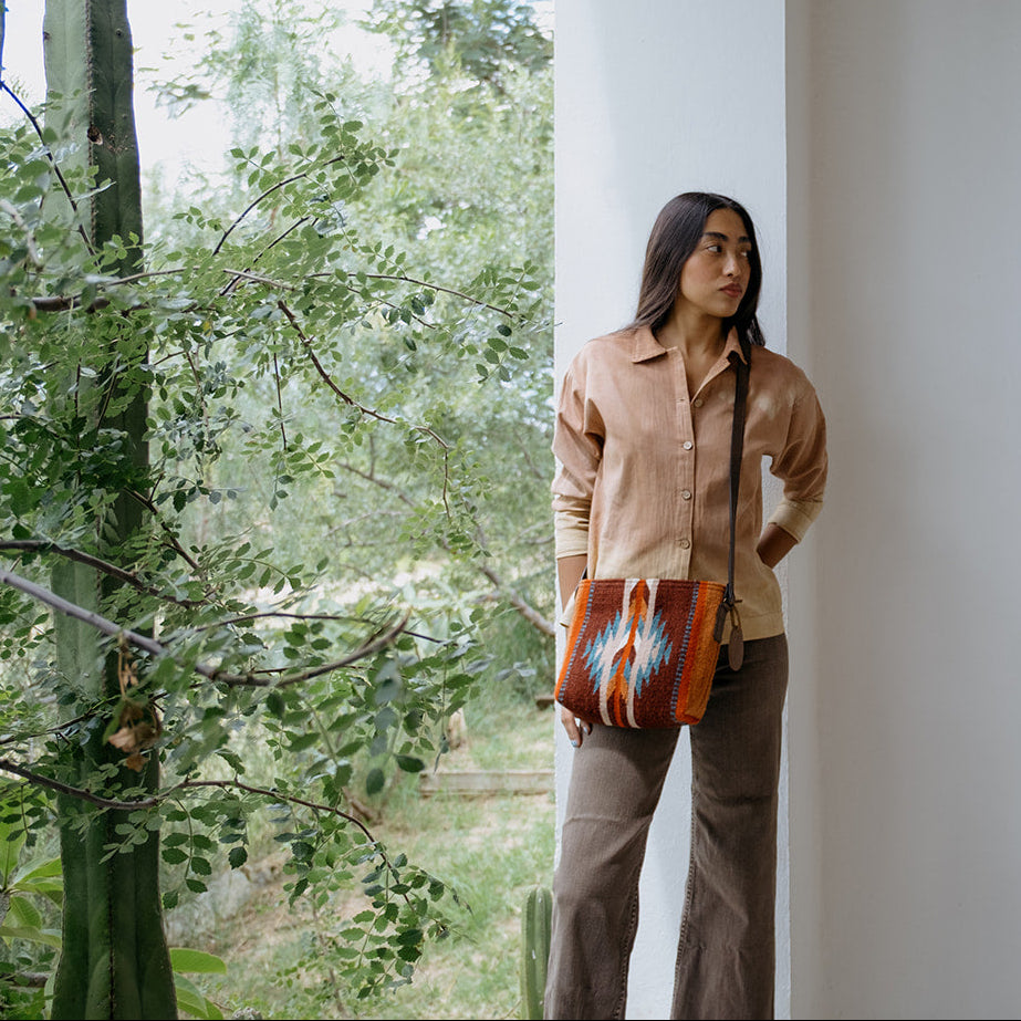 A woman stands in a white architectural corridor holding a brown leather shoulder bag with a colorful geometric pattern, with trees and an outdoor yard visible in the background.