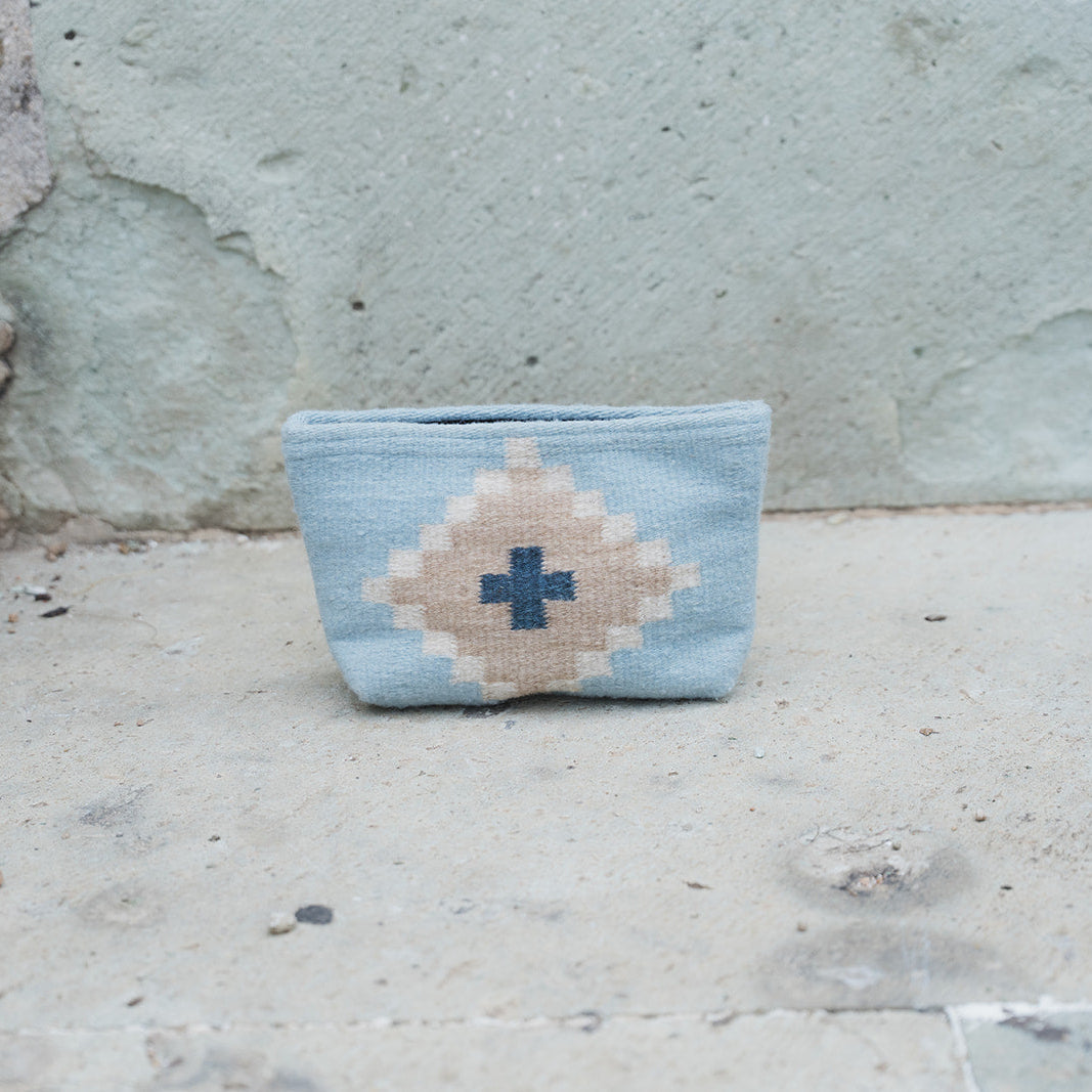 A small woven pouch with a light blue and beige geometric pattern, featuring a dark blue cross in the center, resting on a concrete surface against a textured concrete wall.