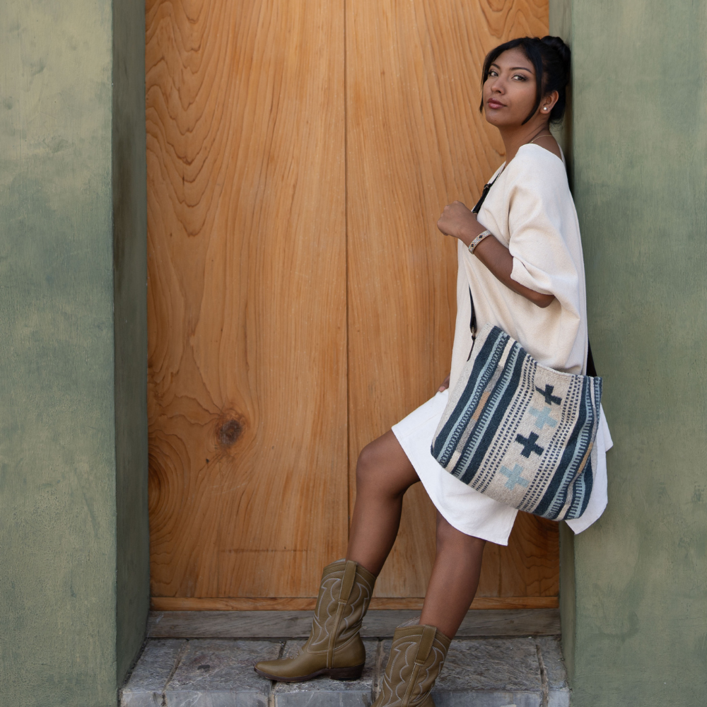 Fashionable woman wearing an AMASOUK Copalita convertible shoulder/crossbody bag made of handwoven wool and leather, featuring blue, cream, honey, and gray tones. Ideal for day-to-night versatility.