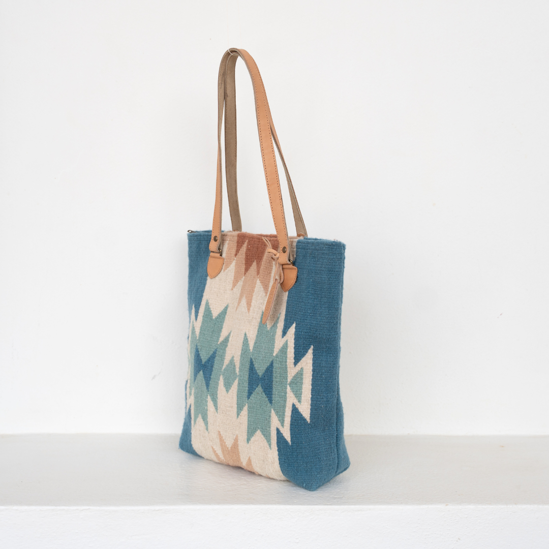 Handwoven bucket bag in rose and blue with abstract diamond and lightning pattern, leather shoulder straps, and relaxed shape, ideal for everyday or beach use.