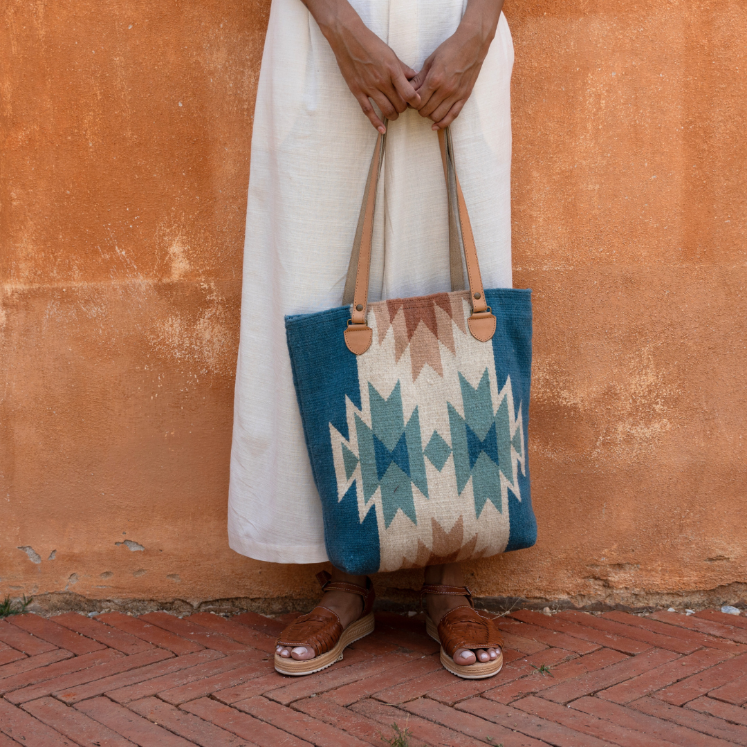 Woman carrying handwoven bucket bag in rose and blue with abstract diamond and lightning pattern, leather shoulder straps, and relaxed shape, ideal for everyday or beach use.