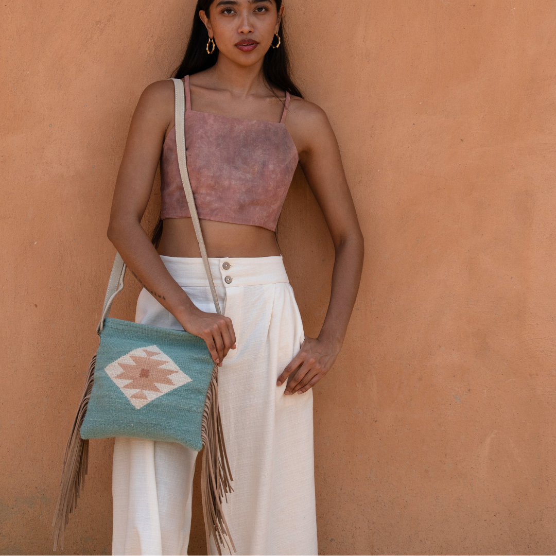Woman carrying handwoven fringe crossbody bag with abstract Zapotec butterfly design in soft blue tones; features flowing fringe, adjustable leather strap, and two internal pockets