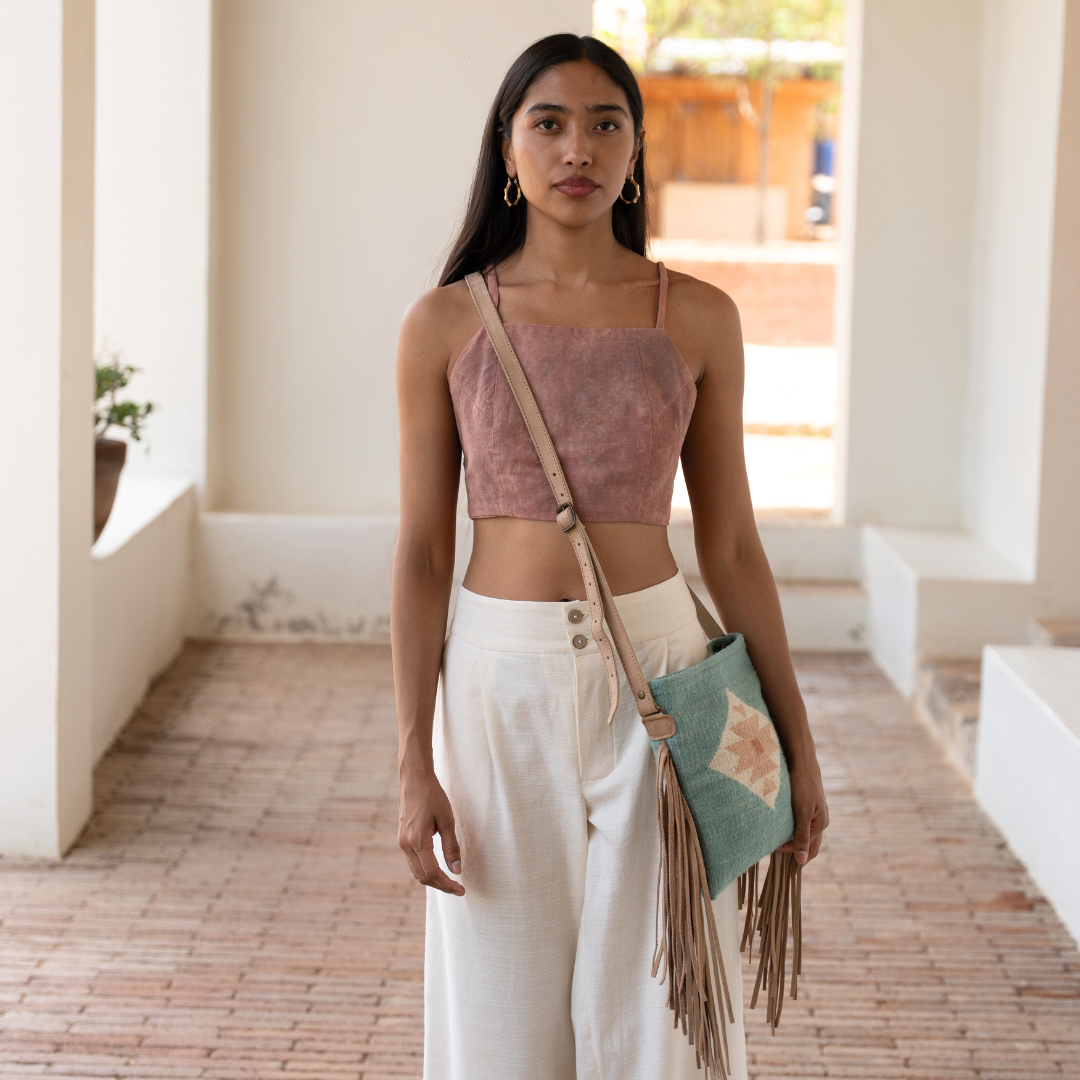 Woman carrying handwoven fringe crossbody bag with abstract Zapotec butterfly design in soft blue tones; features flowing fringe, adjustable leather strap, and two internal pockets