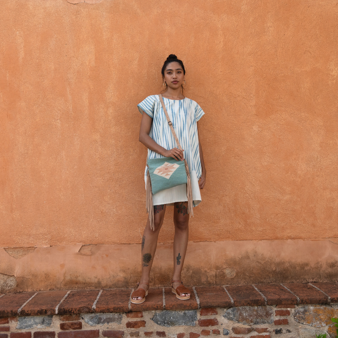 Woman carrying handwoven fringe crossbody bag with abstract Zapotec butterfly design in soft blue tones; features flowing fringe, adjustable leather strap, and two internal pockets