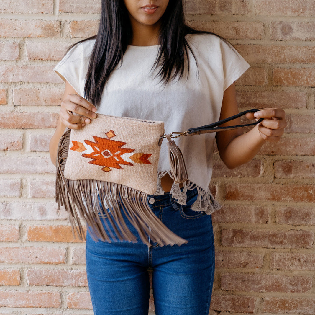 A woman wearing a white t-shirt and blue jeans holds a beige woven clutch bag with orange geometric embroidery and brown fringe against a brick wall background.