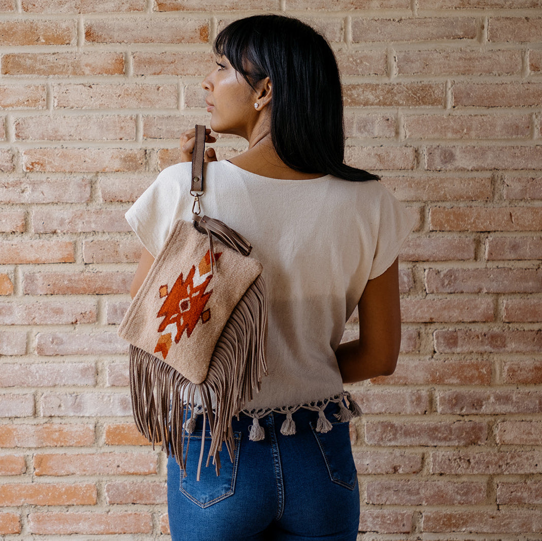 A woman with dark hair is shown from behind, wearing a white t-shirt and blue jeans while carrying a beige fringed shoulder bag with a red geometric pattern, set against a rustic brick wall.