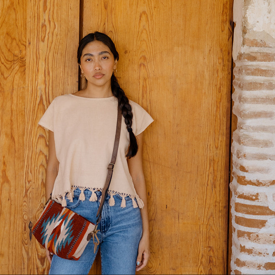 A woman stands in front of a large wooden door wearing a light beige short-sleeve top with fringe trim, blue jeans, and a patterned red, blue, and white crossbody bag.
