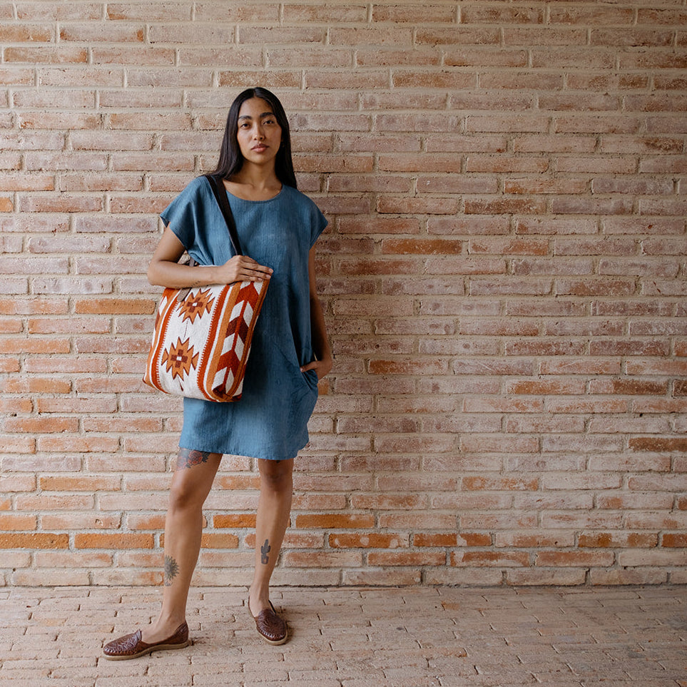 A woman stands against a brick wall wearing a loose blue dress and brown slip-on shoes, carrying a large patterned tote bag with orange, white, and brown geometric designs.