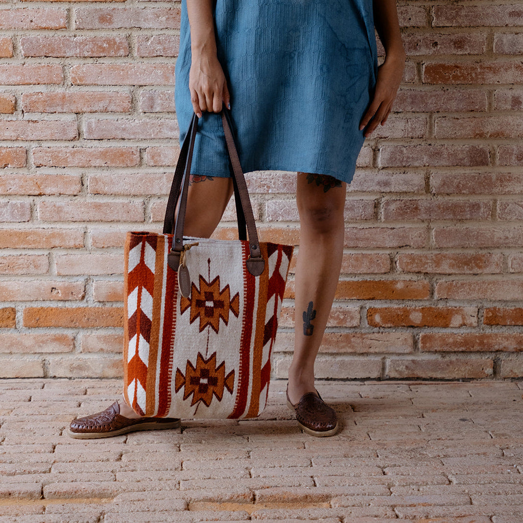 A person wearing a blue dress and brown shoes holds a large woven tote bag with a Southwestern geometric pattern in orange, brown, and white, set against a brick wall background.