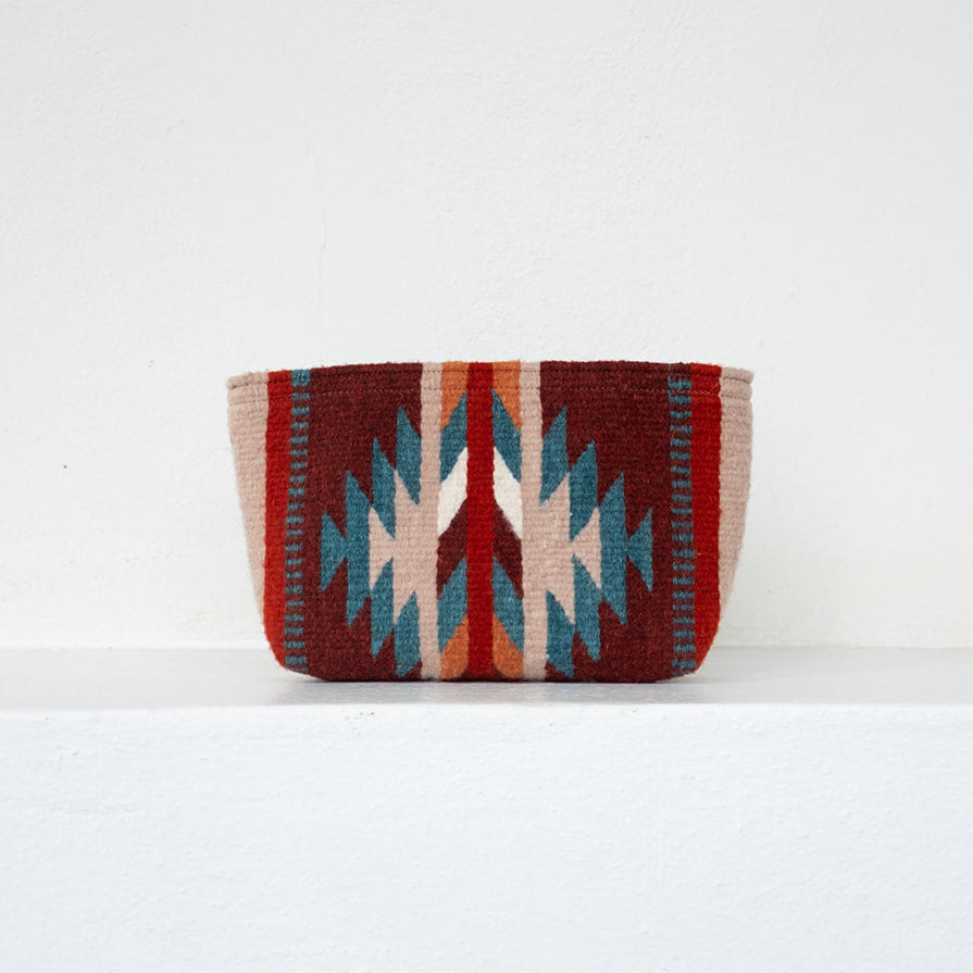 A small woven storage basket with a Southwestern geometric pattern in red, beige, and teal, resting on a white shelf against a white wall.