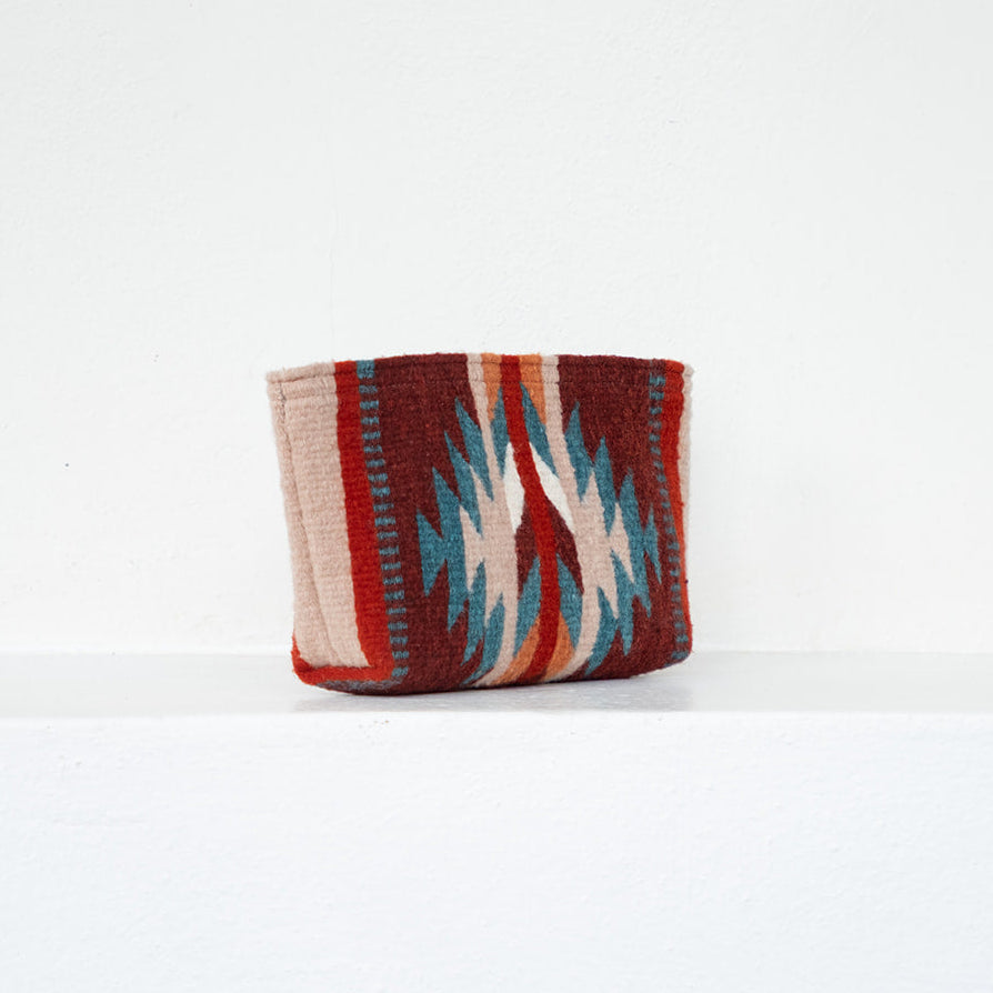 A small woven pouch or wallet with a Southwestern geometric pattern in red, beige, blue, and white, standing on a white shelf against a white wall.