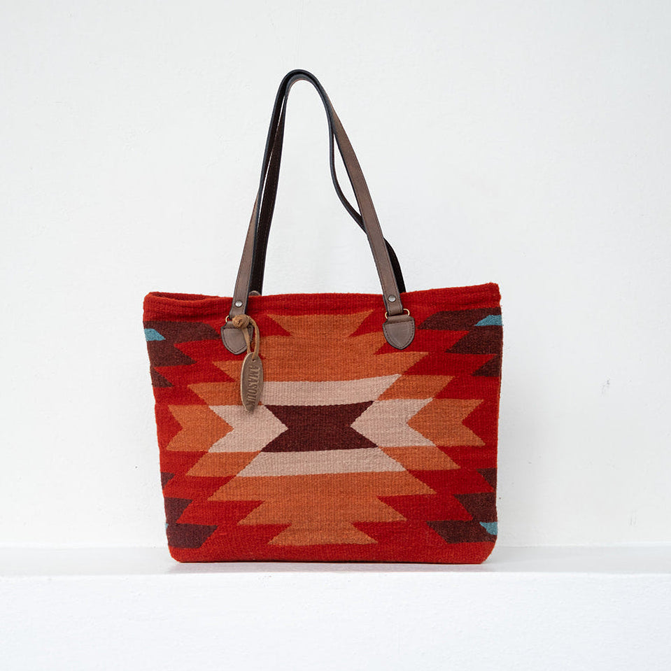 A woven tote bag with a Southwestern geometric pattern in red, orange, beige, and brown, featuring dark brown leather shoulder straps and a hanging leather tag, set against a plain white background.