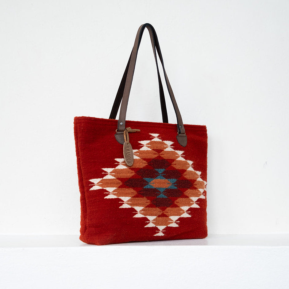 A red woven tote bag with a Southwestern diamond pattern in orange, beige, and blue, featuring dark brown leather shoulder straps, set against a plain white background.