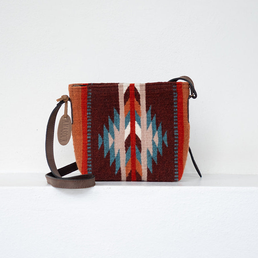 A small woven crossbody bag with a Southwestern geometric pattern in red, orange, brown, cream, and turquoise blue, featuring dark brown leather straps and a hanging leather tag, set against a plain white background.