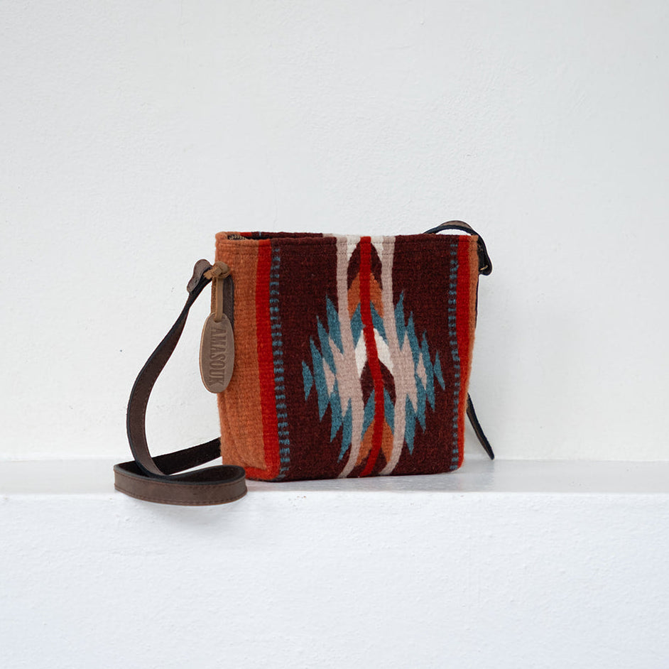 A small woven crossbody bag with a Southwestern geometric pattern in red, brown, cream, and blue, featuring a dark brown leather strap and a hanging leather tag, set against a plain white wall and shelf.