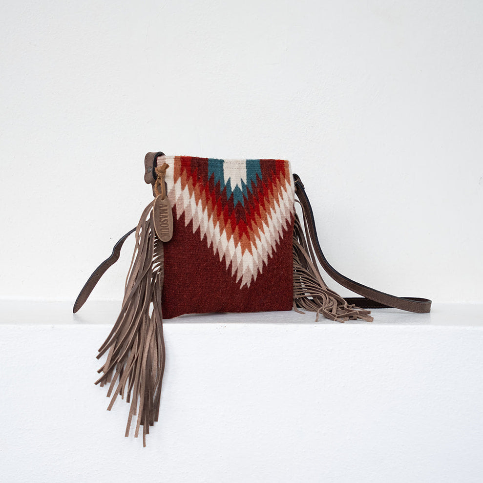 A small fringe clutch bag with a Southwestern-style woven geometric pattern in red, white, and teal, featuring long brown leather fringe and a brown shoulder strap, resting on a white ledge against a white wall.