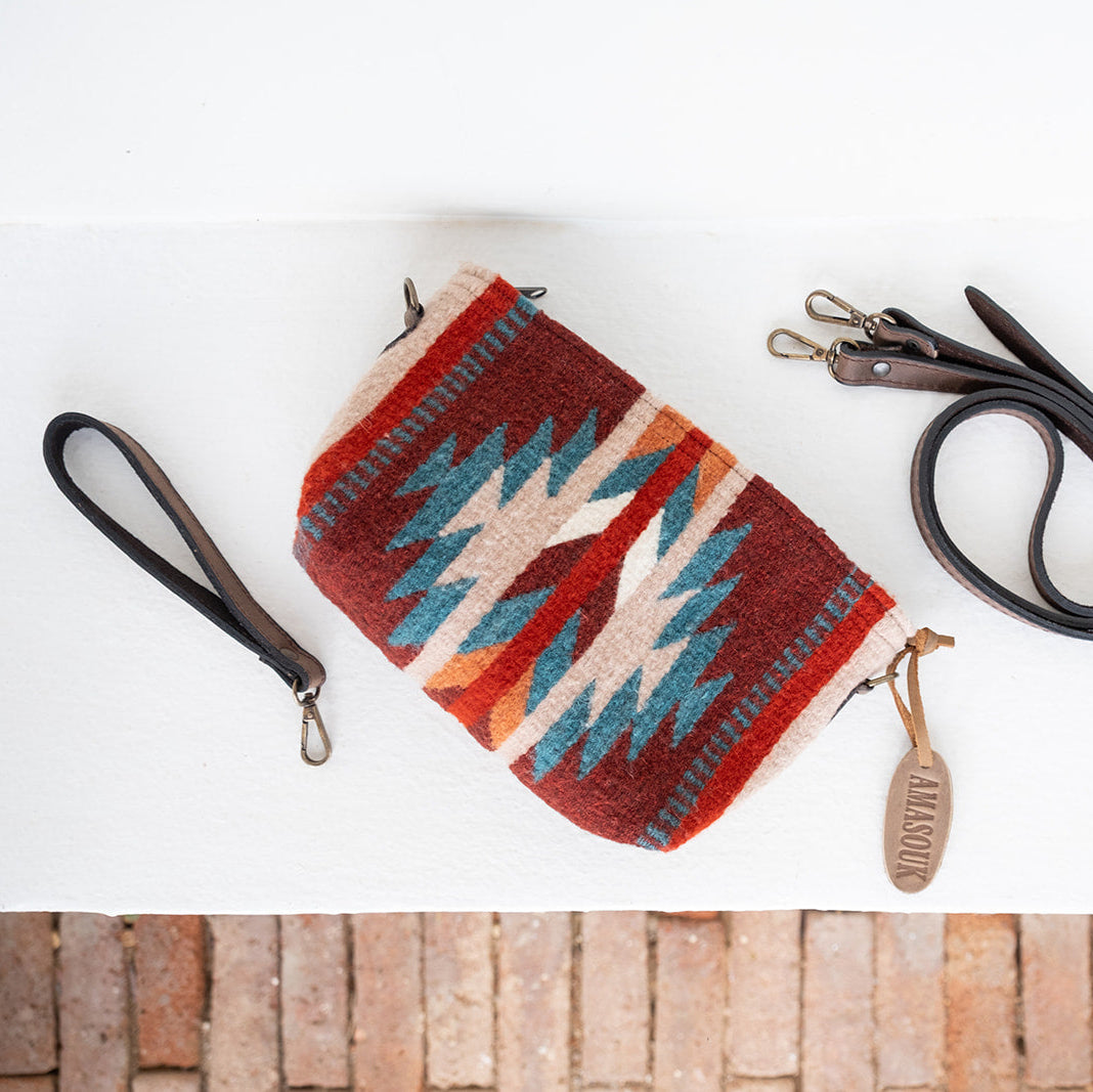 A woven clutch bag with a Southwestern geometric pattern in red, blue, beige, and orange, shown with a black wristlet strap and a black detachable shoulder strap on a white background with a brick pattern at the bottom.