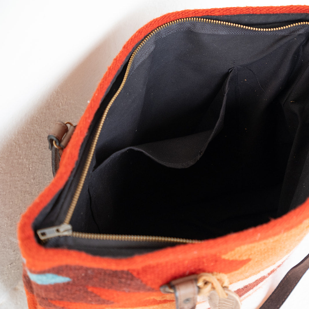 Close-up of the open interior of a bright orange woven bag with a black lining, gold zipper, and brown strap details, photographed against a light neutral background.