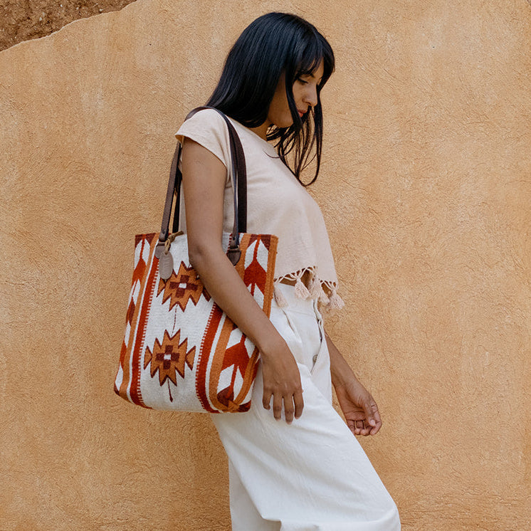 A woman in a white sleeveless top and white wide-leg pants walks against a textured beige stucco wall, carrying a large white tote bag with orange and red geometric patterns and black handles.