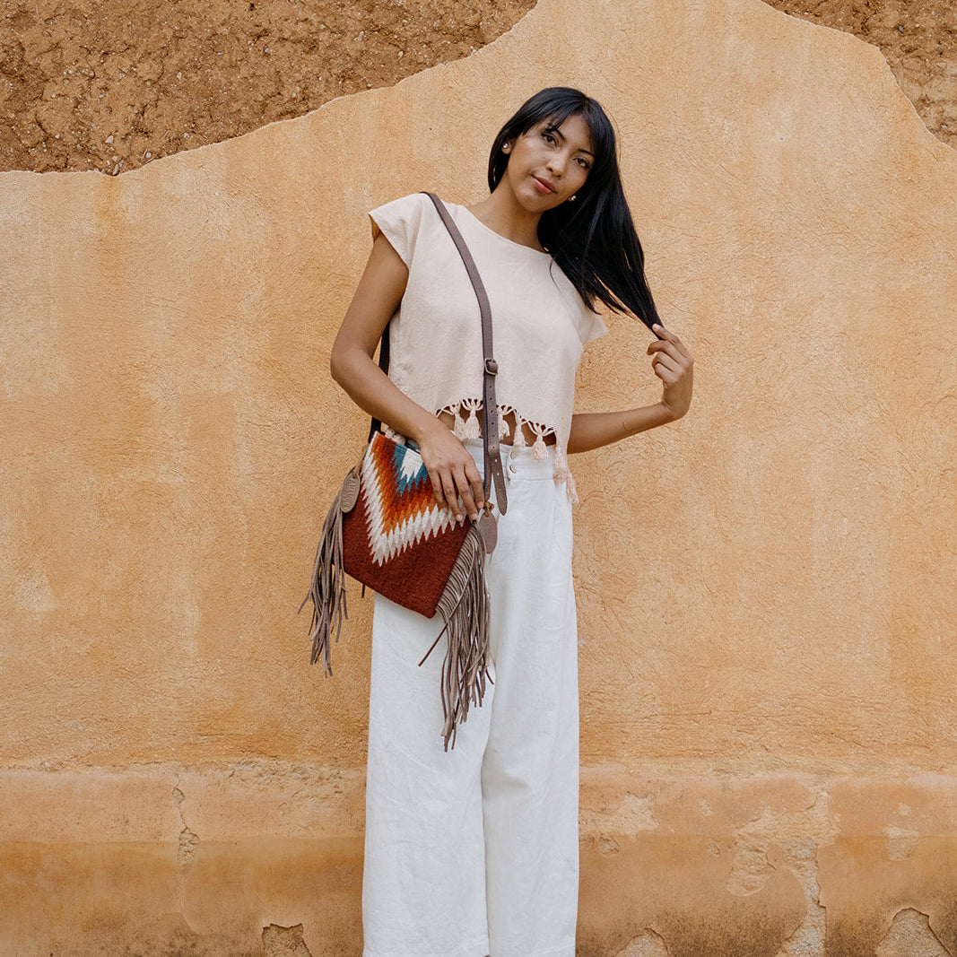 A woman stands against a textured tan stucco wall wearing a cream sleeveless top and white wide-leg pants, carrying a brown leather shoulder bag with fringe and a geometric multicolor woven panel.