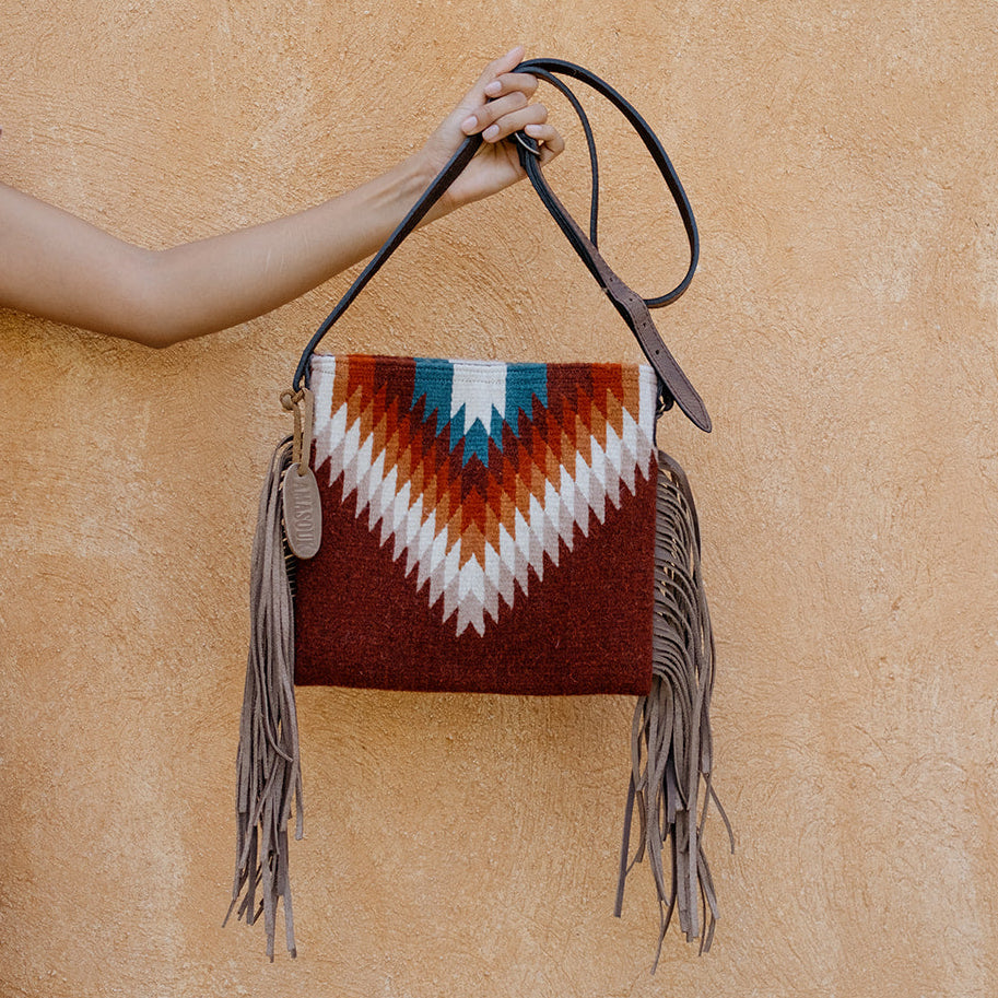 A person in a light pink dress holds up a small woven shoulder bag with a Southwestern-style geometric pattern in rust red, white, and turquoise, featuring long taupe fringe and a dark strap, set against a warm textured stucco wall.