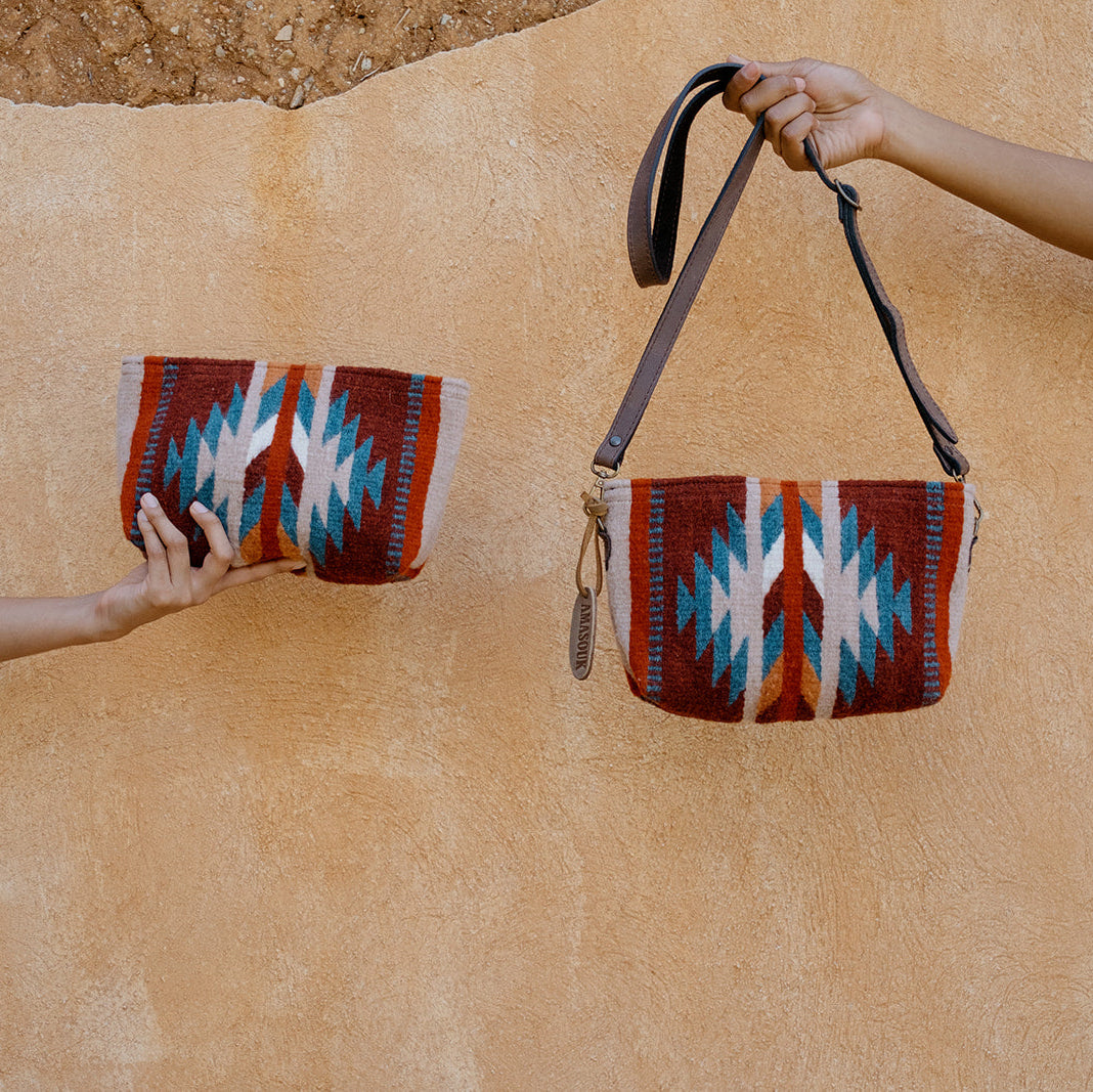 A woven shoulder bag with a Southwestern geometric pattern in red, blue, white, and brown is shown hanging from a person's hand against a textured beige stucco wall. A matching clutch is held by another person's hand on the left.