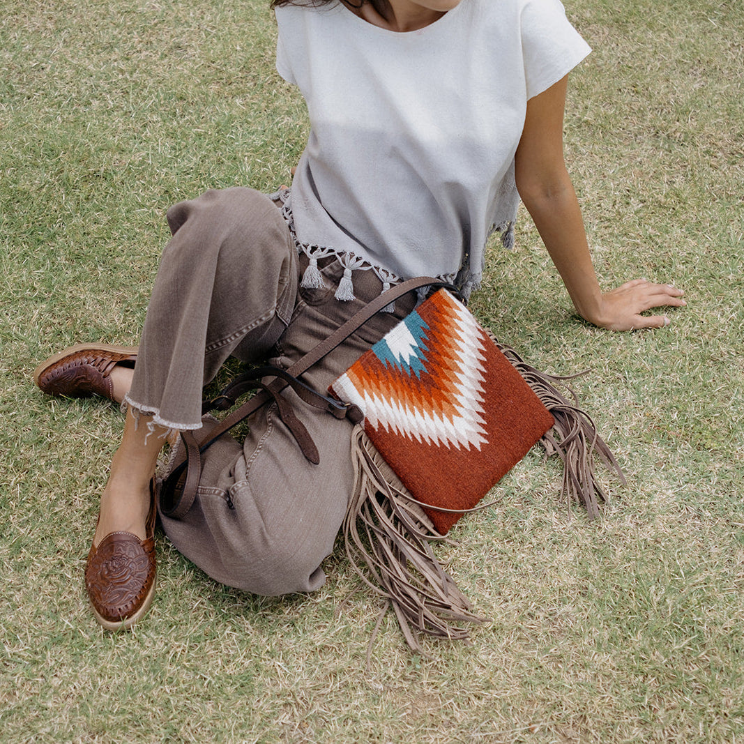 A person sitting on grass wearing a white top and brown pants, with a brown fringed shoulder bag featuring a geometric orange, white, and blue pattern resting against their leg.