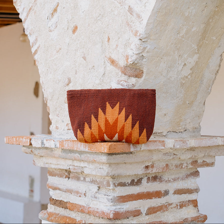 A terracotta-colored woven basket with a geometric orange and yellow sunburst pattern, displayed on a rustic brick and stone pedestal inside a white-walled architectural space.