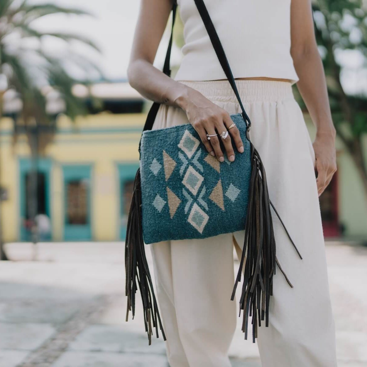 Woman carrying an AMASOUK Jalisco Fringe Crossbody bag with dark brown leather strap and fringe sides, featuring handwoven wool in blue, heather gray, and honey. Festive design with traditional Zapotec patterns.

