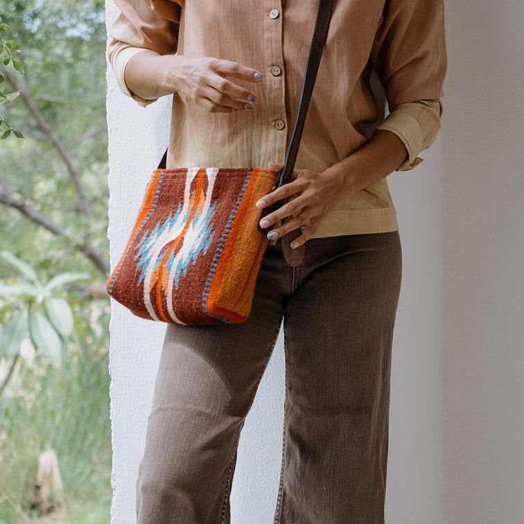 A person wearing brown wide-leg pants and a tan button-up shirt carries a woven orange, red, and blue striped shoulder bag while standing next to a white wall with green foliage on the left.